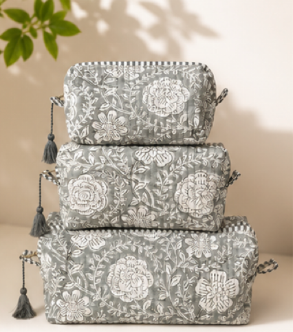 Three stacked gray fabric pouches with white floral embroidery and black tassels, set against a warm beige background with soft leaf shadows.