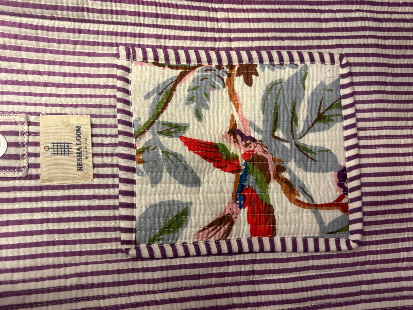 A close-up of a quilted fabric product by Resha Loom, featuring a central patchwork design with a colorful hummingbird and tropical leaves on a white background, surrounded by a purple and white striped border.