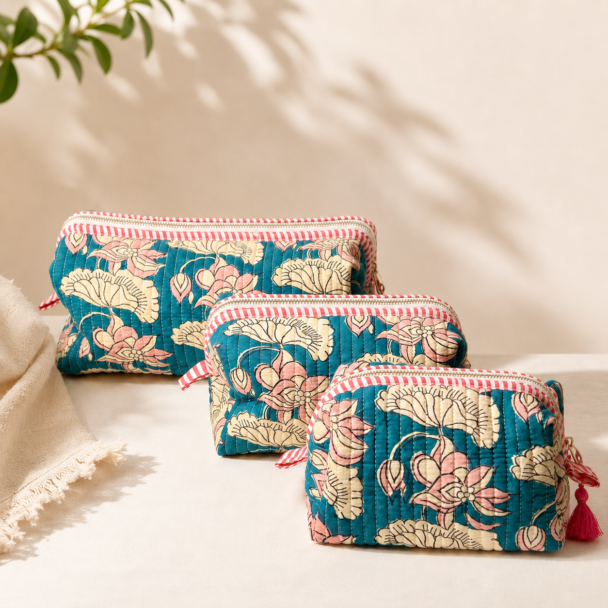 Three quilted zip pouches with a teal lotus floral print, pink and cream stripes, and red tassels, arranged diagonally on a light beige surface with soft shadows.