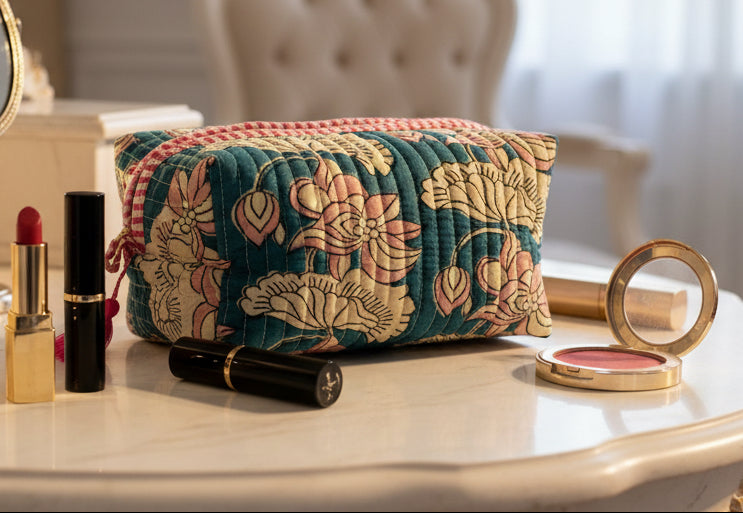 A rectangular quilted cosmetic pouch with a floral print in teal, pink, and cream, featuring a red-and-white checkered top trim and pink tassels on the sides, set against a plain light gray background.