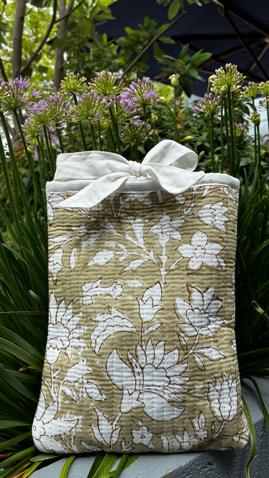 A quilted fabric pouch with a white floral print and a large white bow, displayed outdoors among green plants and purple flowers.
