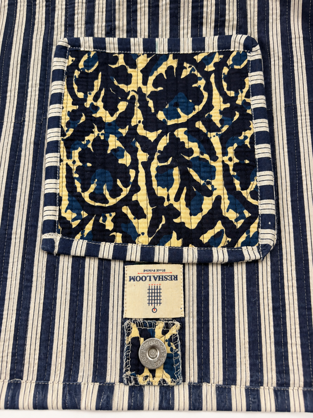 Close-up of a Reshloom fabric product showing a navy and cream striped border, a navy and cream damask patterned pocket, and label with a metal snap.