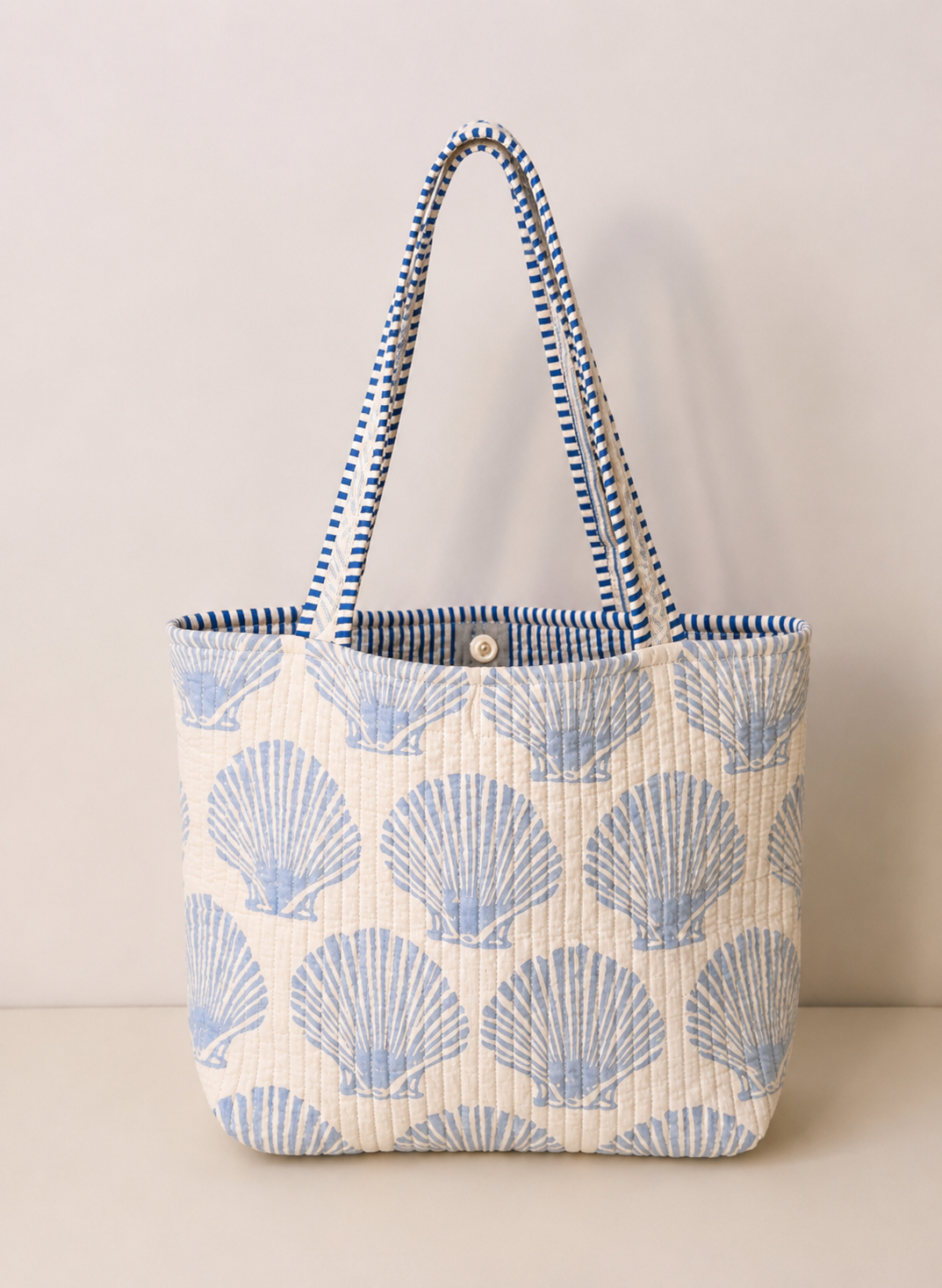 A quilted tote bag with a blue scallop shell pattern on a cream background, featuring blue and white striped woven shoulder straps and a small gold button closure at the top.
