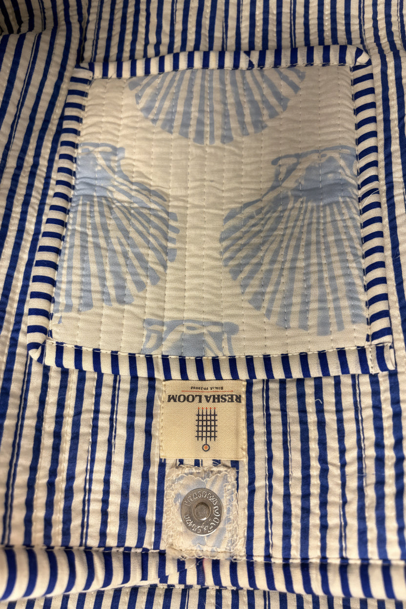 Close-up of a quilted baby blanket featuring a blue and white striped border, a central cream patch with a light blue scallop shell pattern.