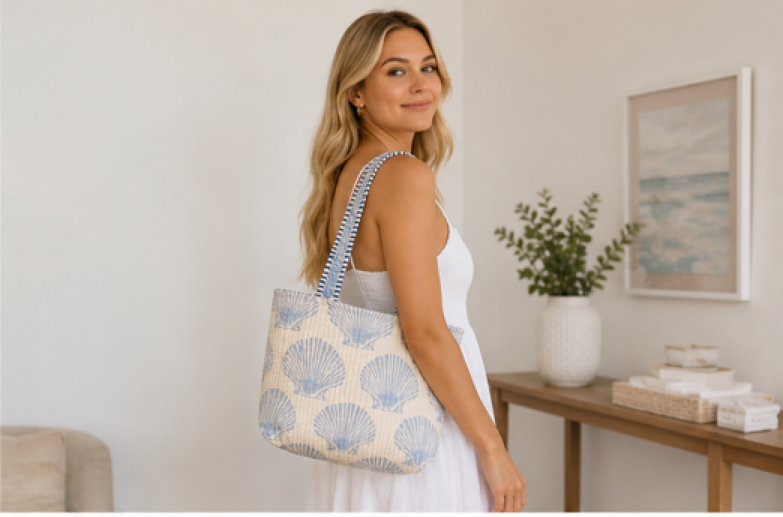 A woman in a white dress carries a cream canvas tote bag with a blue scallop shell pattern and a striped shoulder strap in a bright, modern room.