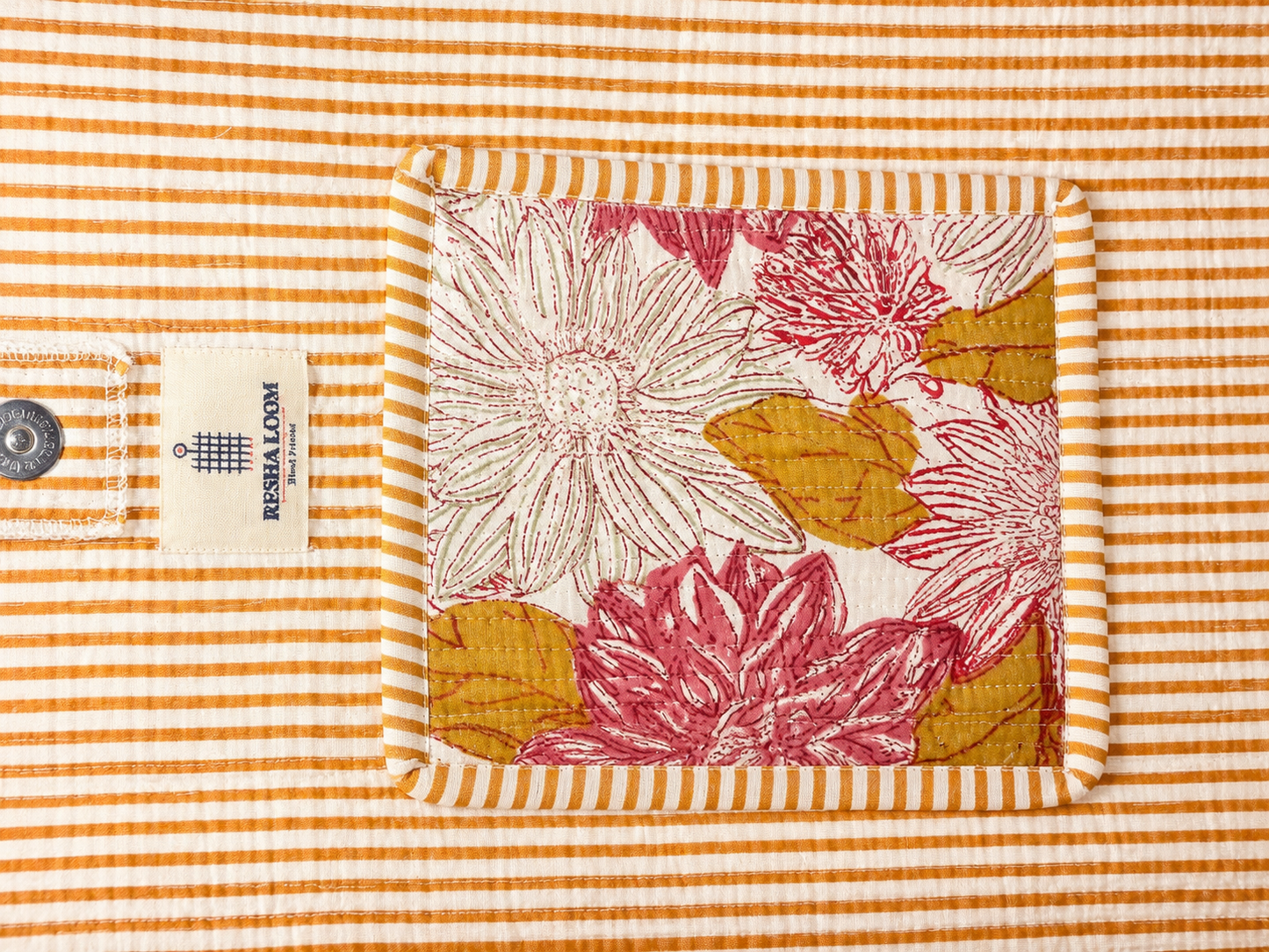 A close-up of a quilted fabric bag, featuring a central floral compartment design with pink and mustard yellow flowers on a white background, surrounded by a striped border. 