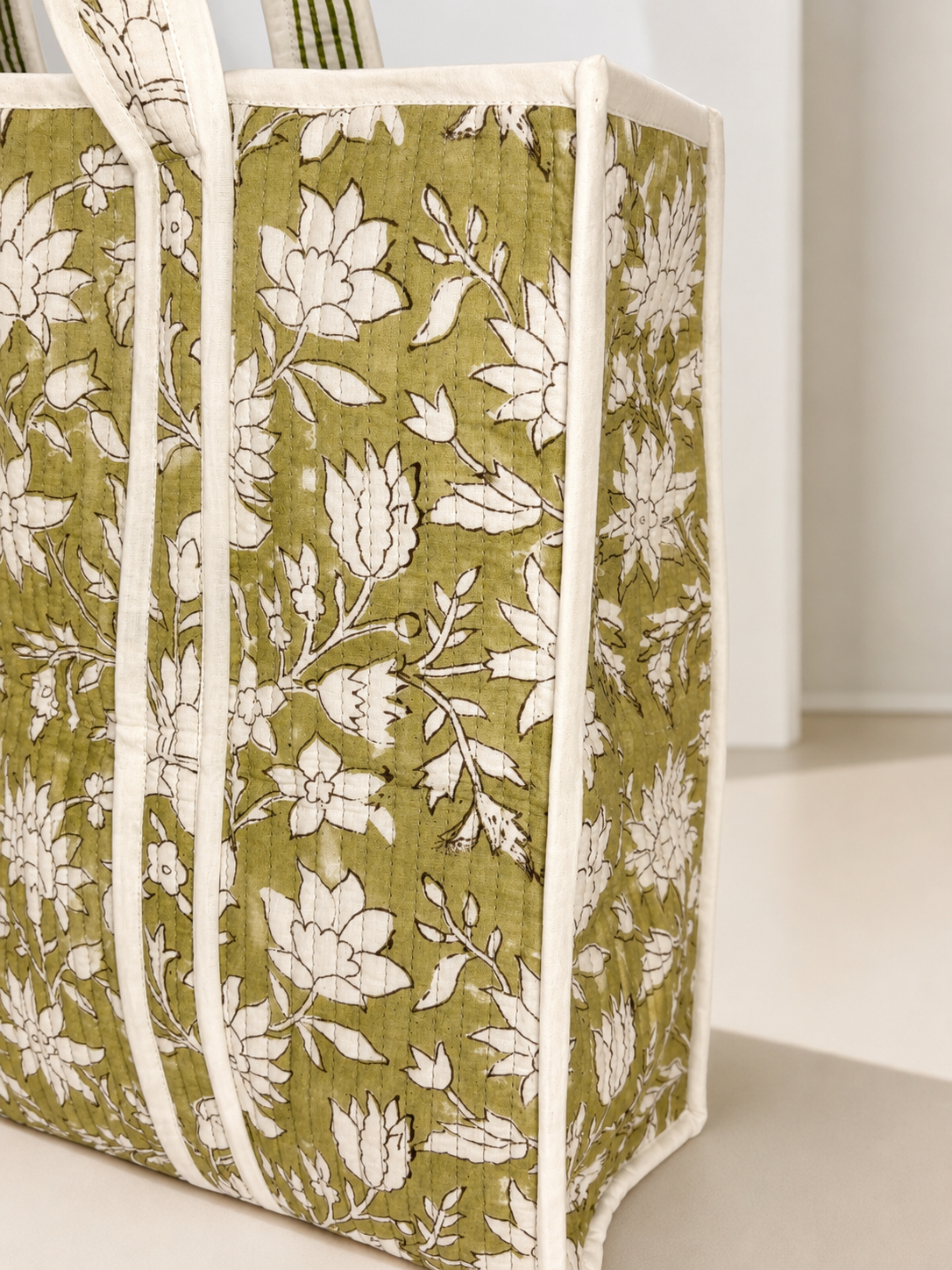 A close-up of a quilted fabric tote bag with an olive green and white floral pattern, featuring cream-colored piping and handles.