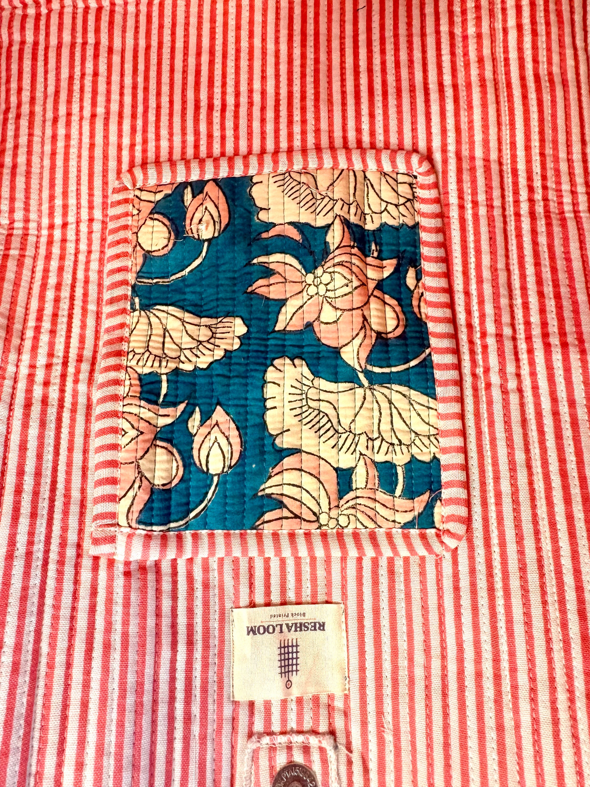 A close-up of a quilted textile product, likely a kantha blanket or quilt, featuring a red and white striped body with a central square patch of teal blue fabric printed with pink and beige lotus flowers. 
