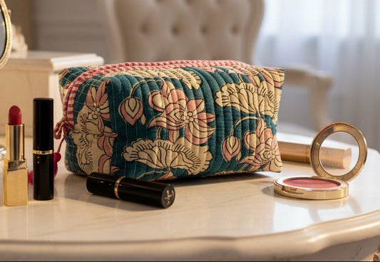A rectangular quilted cosmetic pouch with a floral print in teal, pink, and cream, featuring a red-and-white checkered top trim and pink tassels on the sides, set against a plain light gray background.