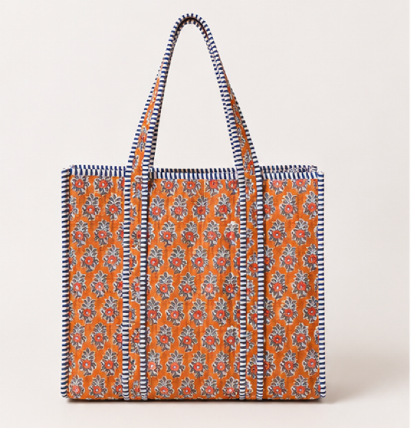 A tote bag with a warm orange base, covered in a detailed multicolor floral print, and finished with a navy and cream striped border and matching woven shoulder straps.