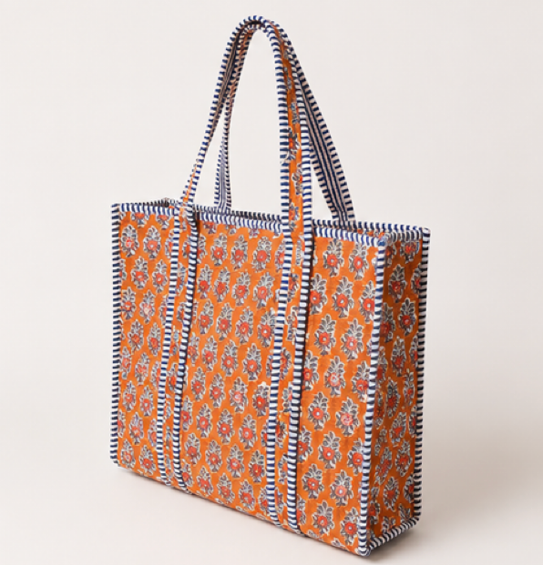 A tote bag with a warm orange base, covered in a detailed multicolor floral print, and finished with a navy and cream striped border and matching woven shoulder straps.